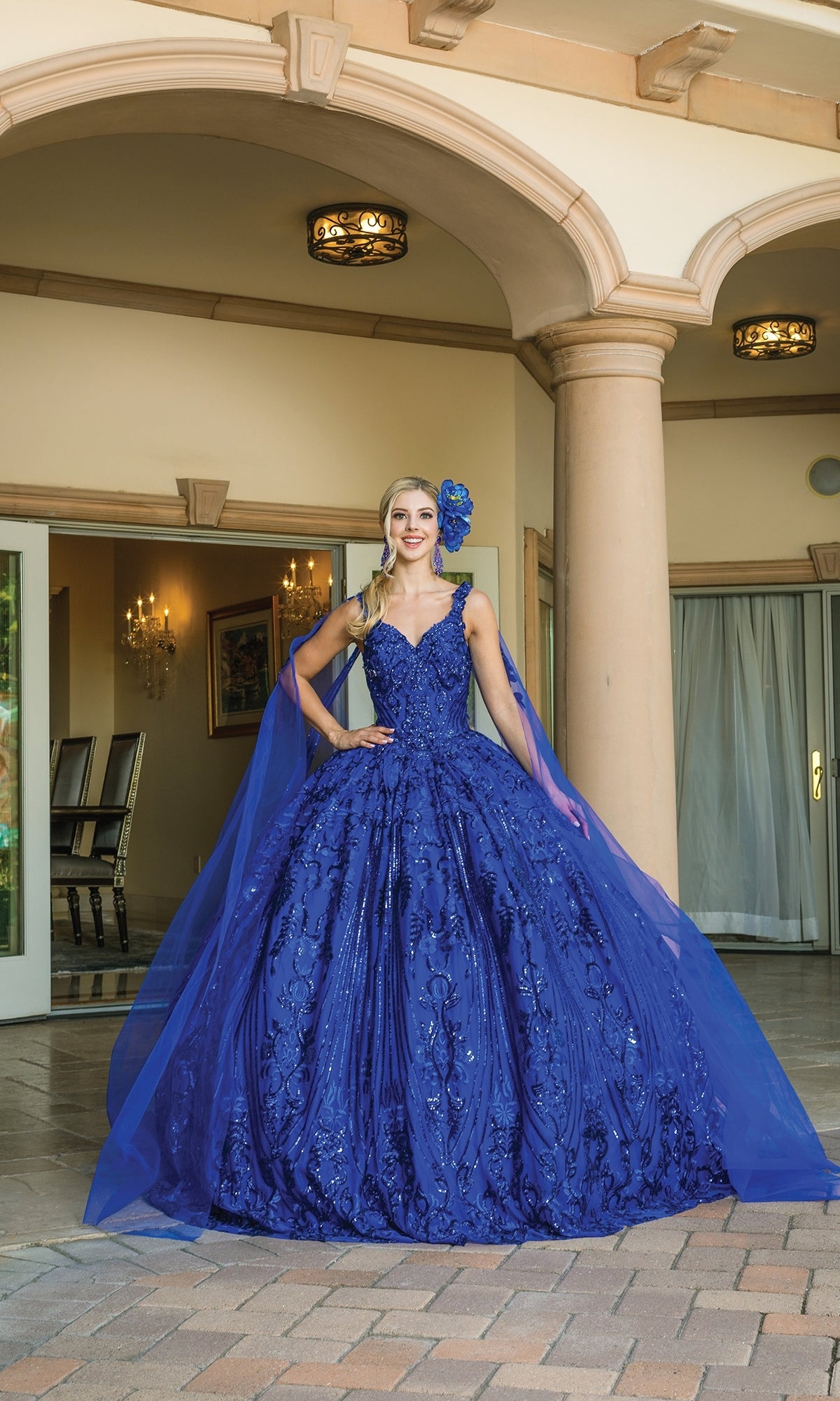 A woman poses outdoors in front of columns, wearing the Quinceanera Dress 1649 By Dancing Queen—a royal blue ball gown with a sweetheart neckline, tulle cape, and matching headpiece.